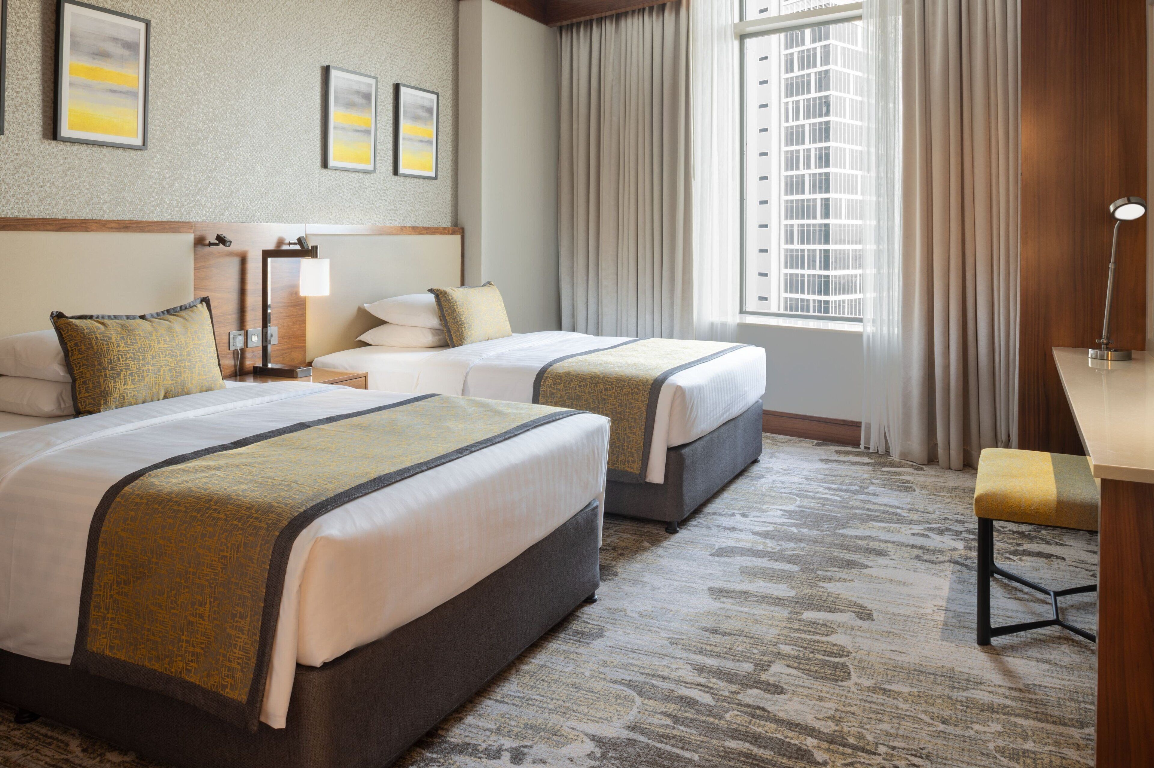 A hotel room with two beds, a desk, a chair, a lamp, and a window with curtains.