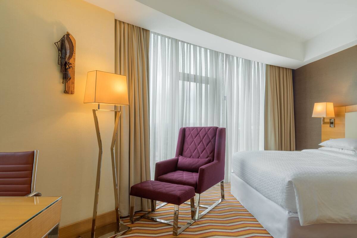 A hotel room with a bed, a purple armchair, a side table, a floor lamp, and a large window with curtains.