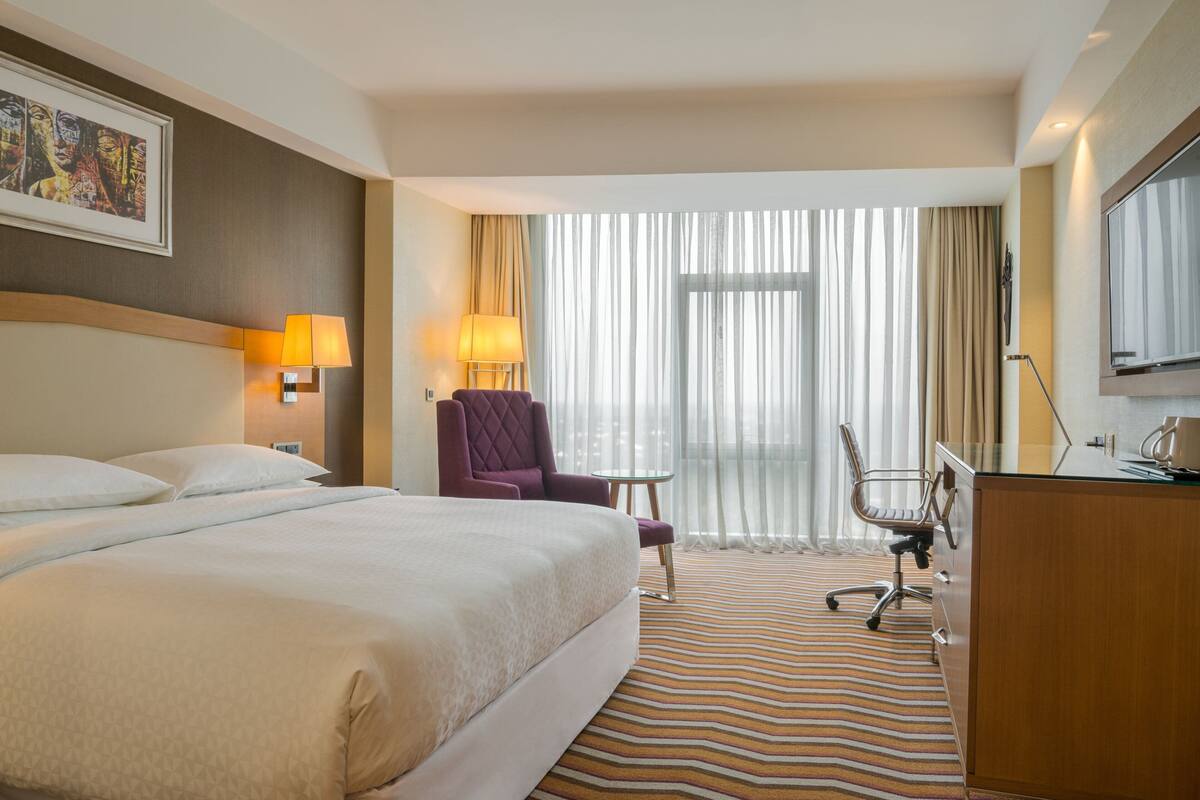 A hotel room with a large bed, a desk, a chair, a lamp, and a large window with curtains.