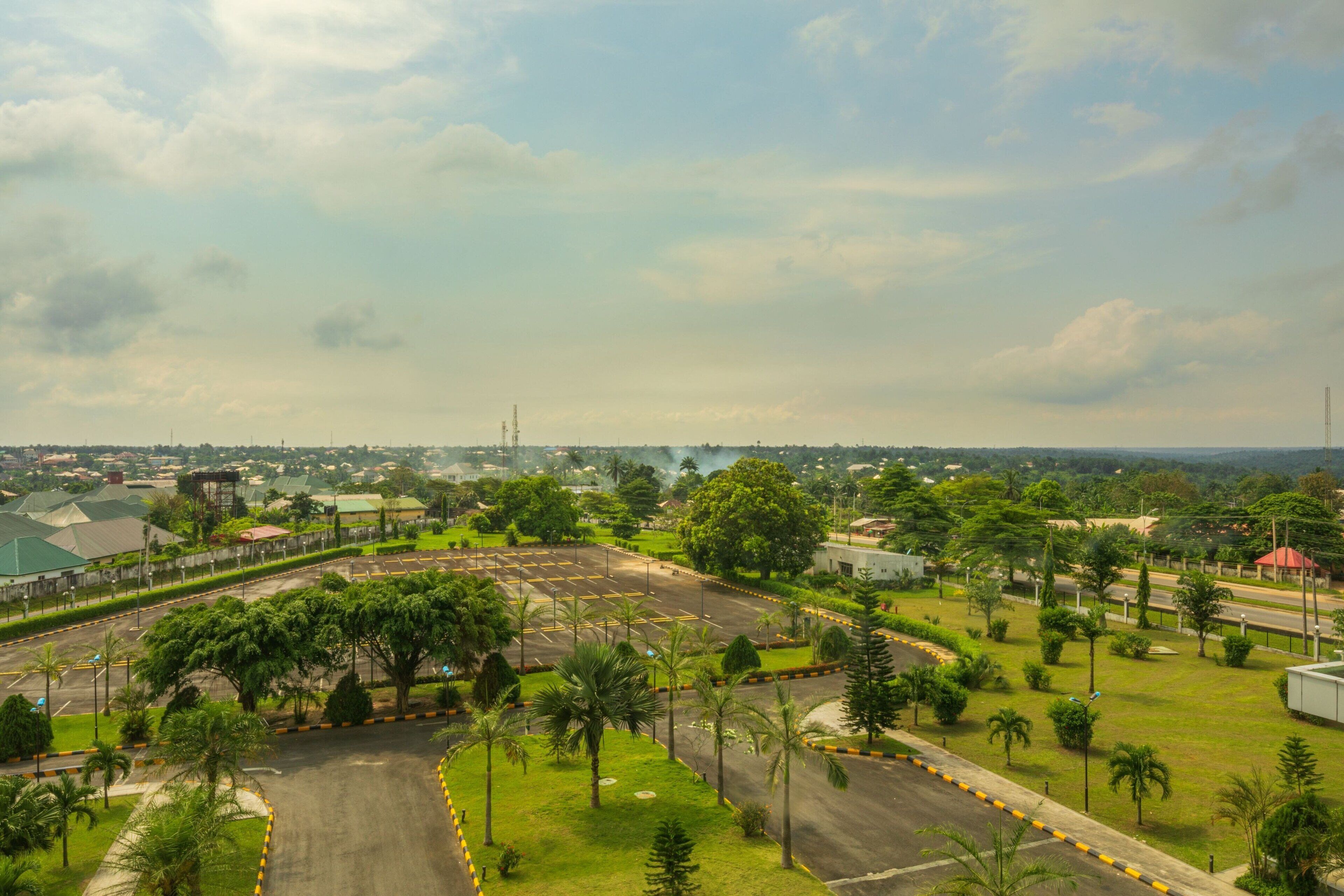 A large parking lot with multiple rows of parking spaces, surrounded by greenery and trees.
