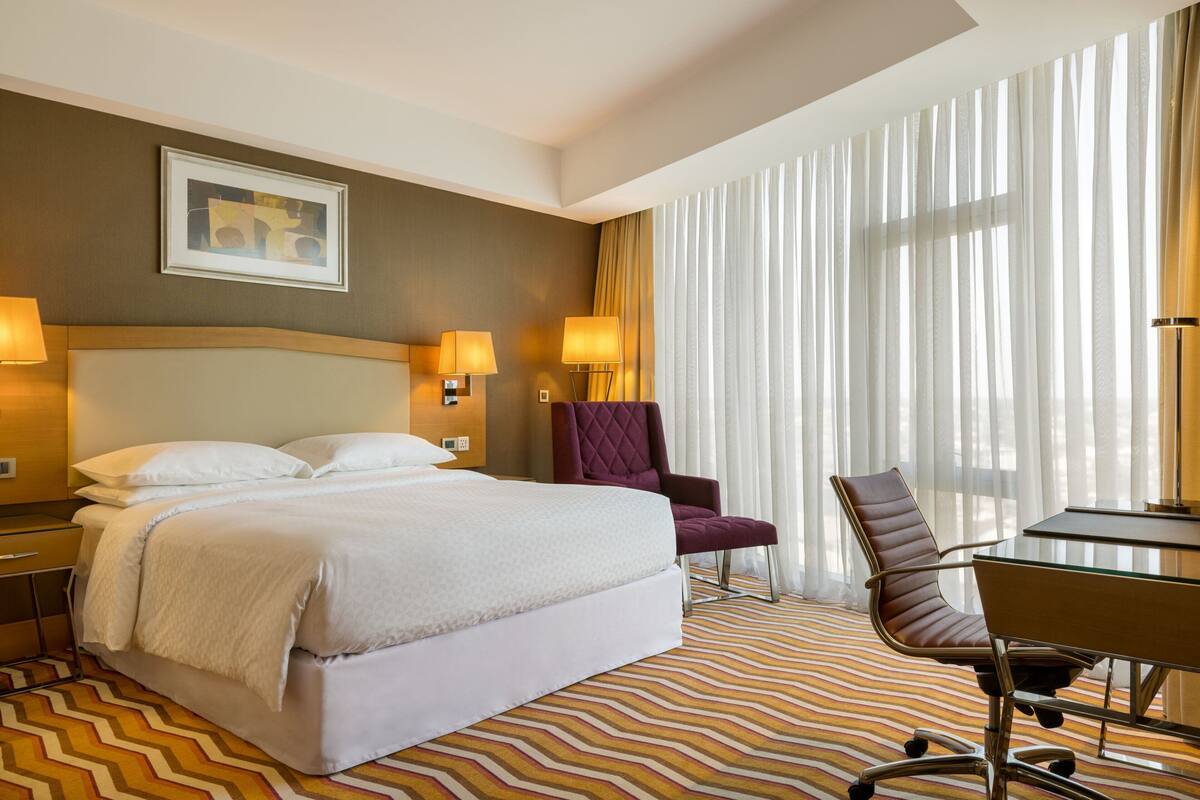 A hotel room with a large bed, two bedside lamps, a desk with a chair, and a window with sheer curtains.