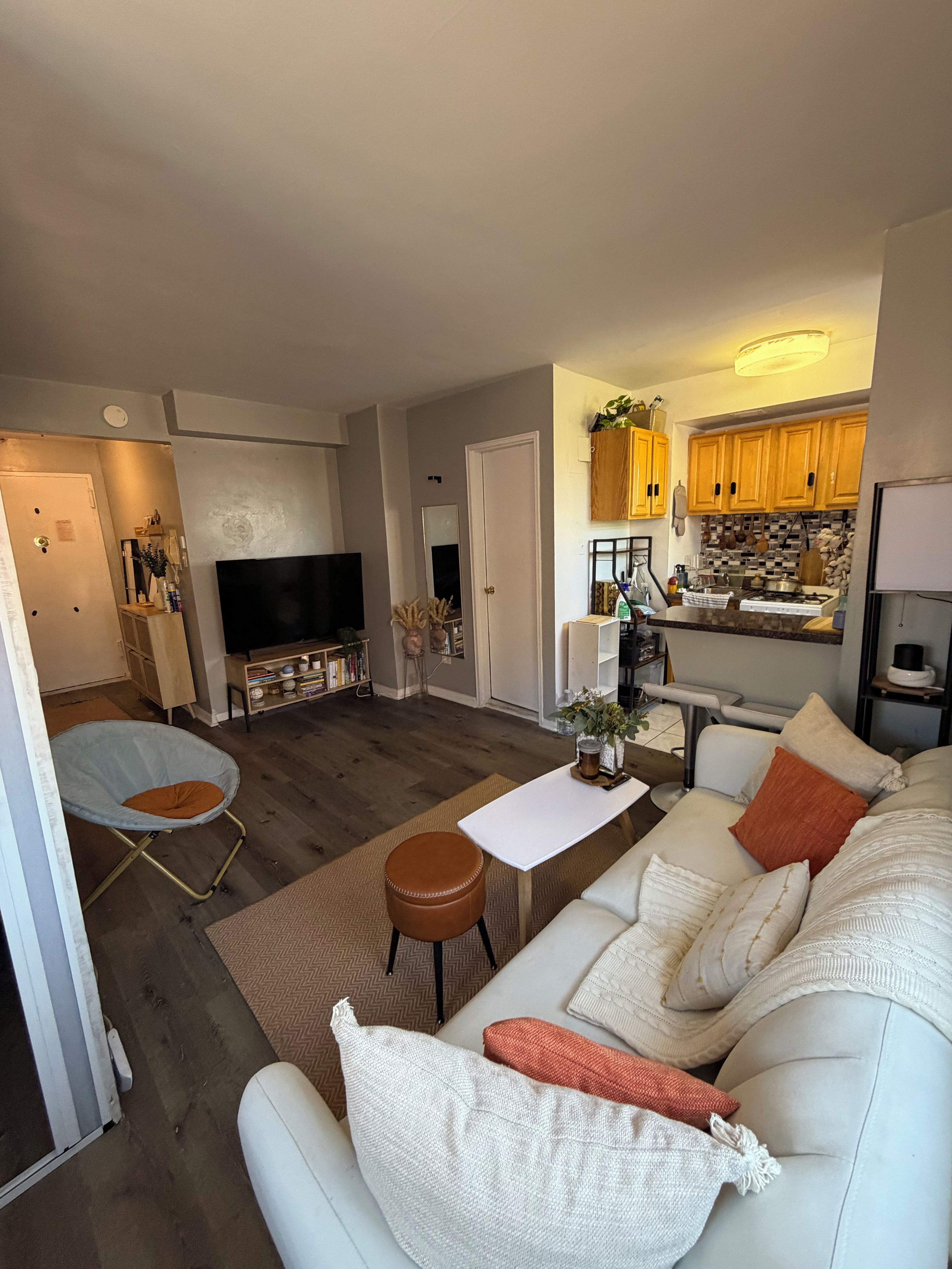 A living room with a sofa, a coffee table, and a TV. There is a kitchen in the background with wooden cabinets and a tiled backsplash.