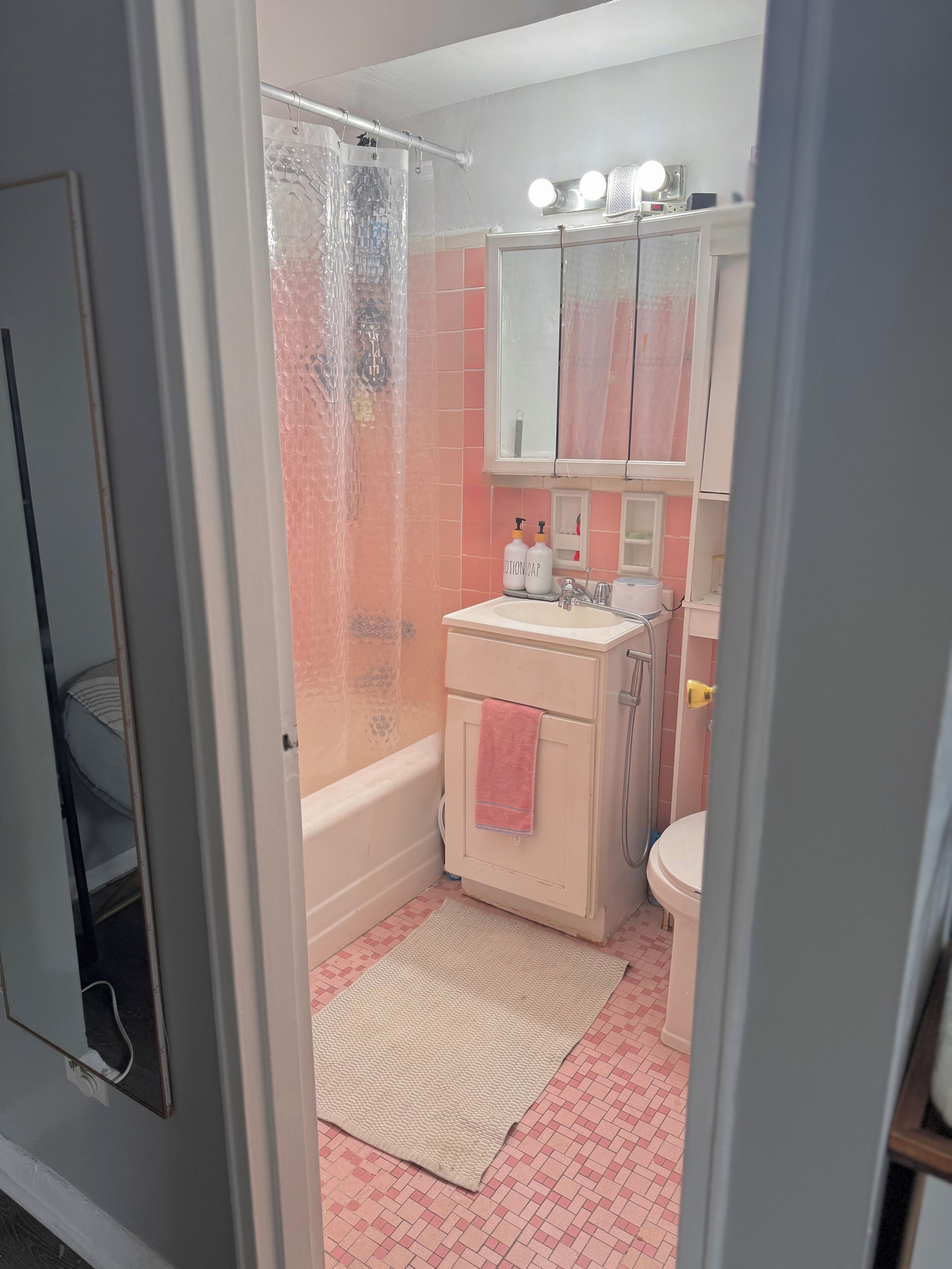 A bathroom with a bathtub, toilet, and sink. There is a shower curtain, a mirror with lights, and a towel hanging on the door.