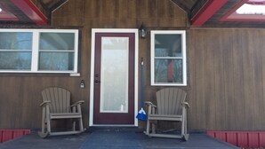 Un edificio de madera con puerta roja, dos ventanas y dos sillas Adirondack en el porche.