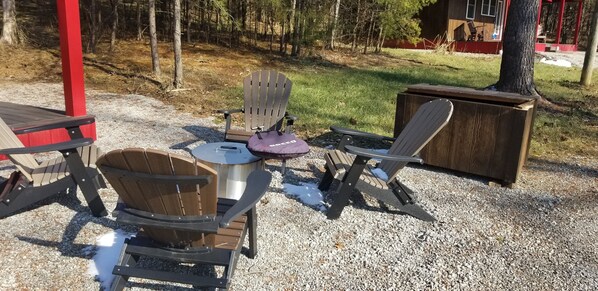 A set of Adirondack chairs and a metal table with a bag on it, set on a gravel surface with a wooden structure in the background.