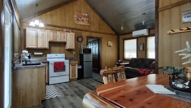 Una cocina acogedora con armarios de madera, una estufa y un refrigerador. Hay una mesa de comedor con sillas y una ventana con persianas.