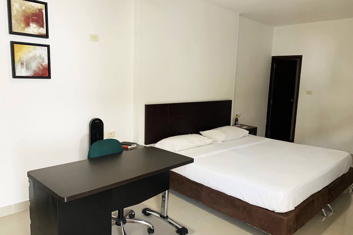 A hotel room with a bed, a desk, and two framed artworks on the wall.