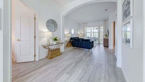 Un couloir avec une porte blanche, un miroir rond, un canapé bleu et une table à manger agrémentée d’un vase.