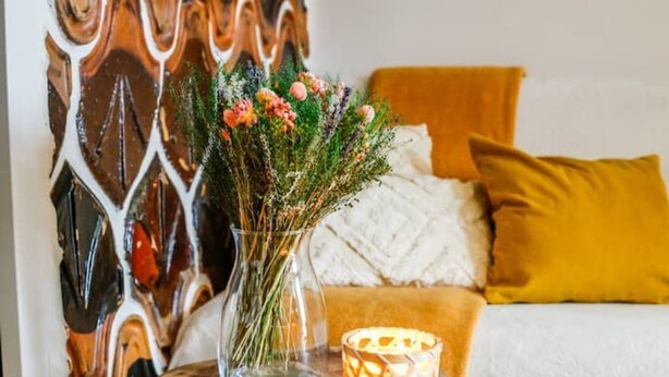 Una habitación con una mesita de madera, un florero con flores, una vela encendida y un sofá con un cojín amarillo.