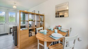 Un coin repas avec une table en bois dressée pour quatre personnes, entourée de chaises blanches. Une bibliothèque en bois, agrémentée de rideaux, se trouve à gauche, et on aperçoit un espace cuisine à travers une porte ouverte.