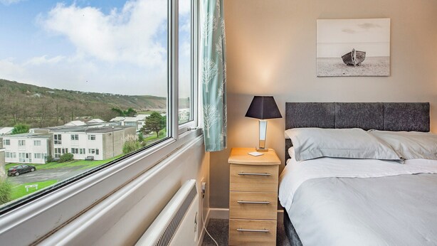 Une chambre d’hôtel avec une grande fenêtre, un lit, une table de chevet avec une lampe, une commode et un tableau représentant un bateau accroché au mur.
