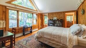 A bedroom with a large bed, wooden walls, and large windows offering a view of the forest. - Rustic and luxurious 4 bedroom house on secluded NH lake! (Pittsfield)