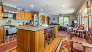 A spacious kitchen with wooden cabinets, a large island, and a dining area with wooden chairs and a rug. - Rustic and luxurious 4 bedroom house on secluded NH lake! (Pittsfield)