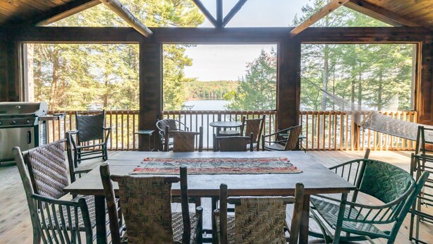 A covered outdoor dining area with a table and chairs, a grill, and a view of a forested area. - Rustic and luxurious 4 bedroom house on secluded NH lake! (Pittsfield)