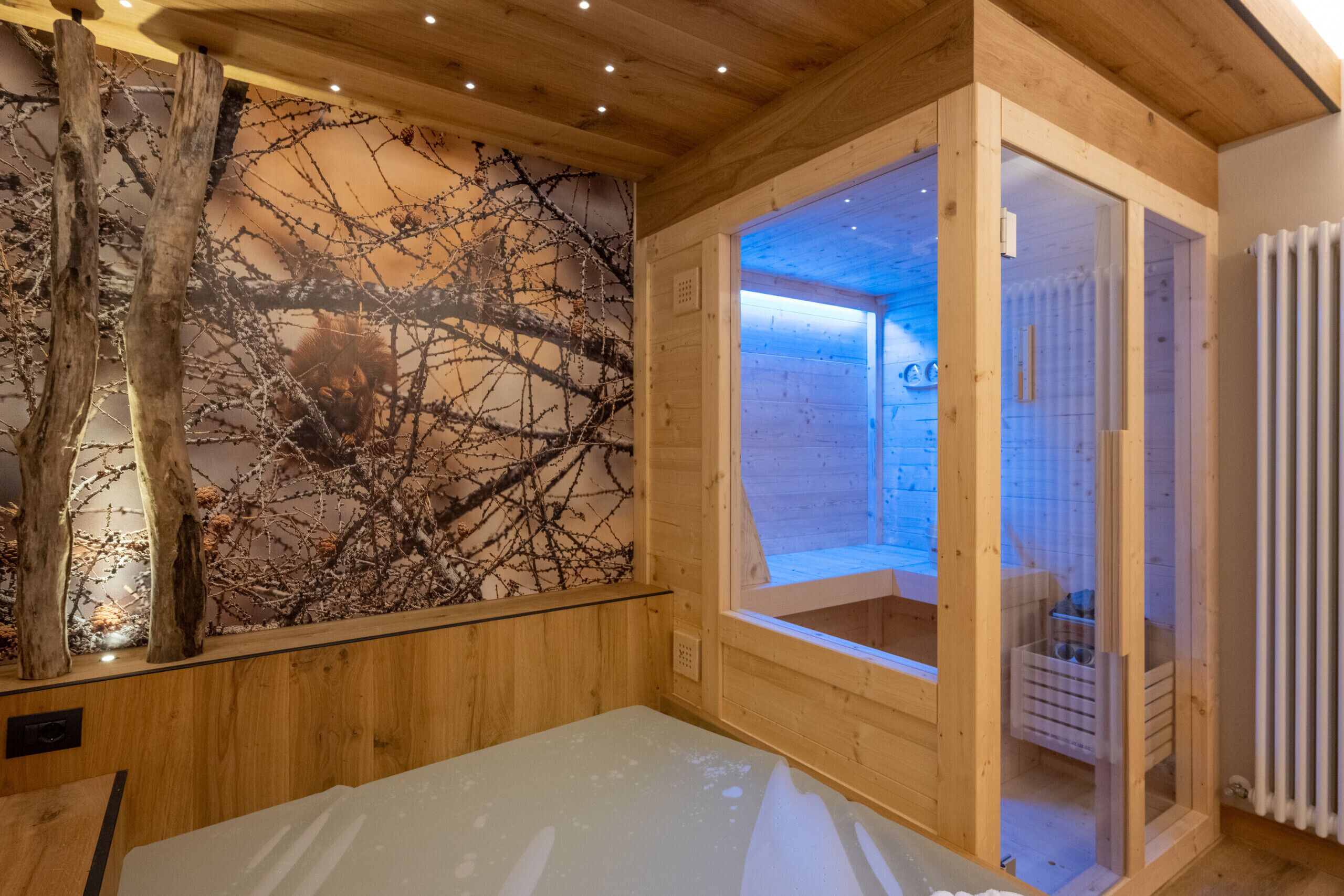 Innenansicht einer Holzhütte mit Sauna, einer Holzwand mit Baumzweig-Fresko und Blick ins Freie.