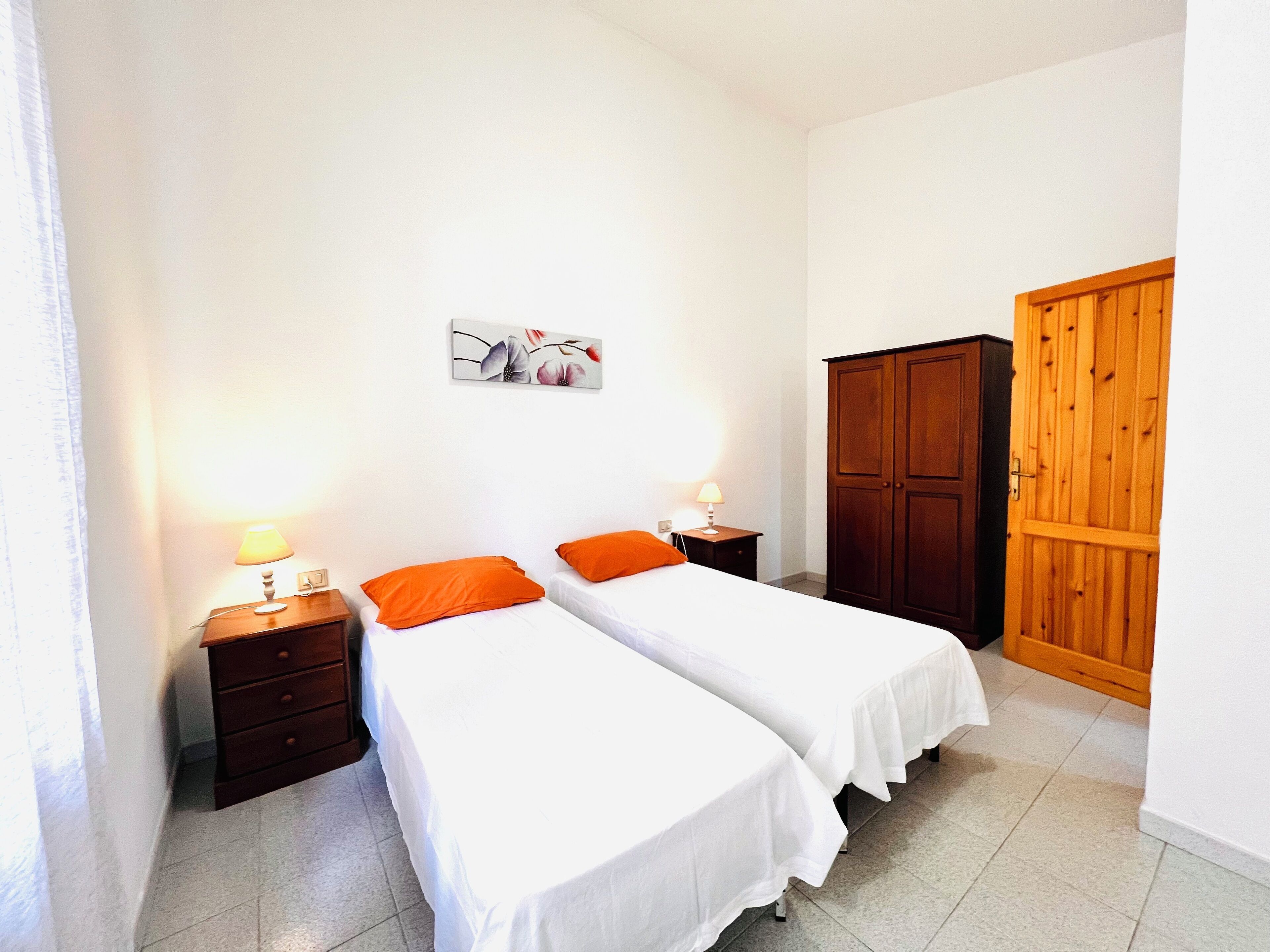 A hotel room with two single beds, a wooden wardrobe, a nightstand with a lamp, and a floral artwork on the wall.