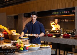 Un chef prépare des plats dans une cuisine moderne au décor en bois, avec un buffet proposant divers mets et un poste de travail équipé pour la préparation culinaire.