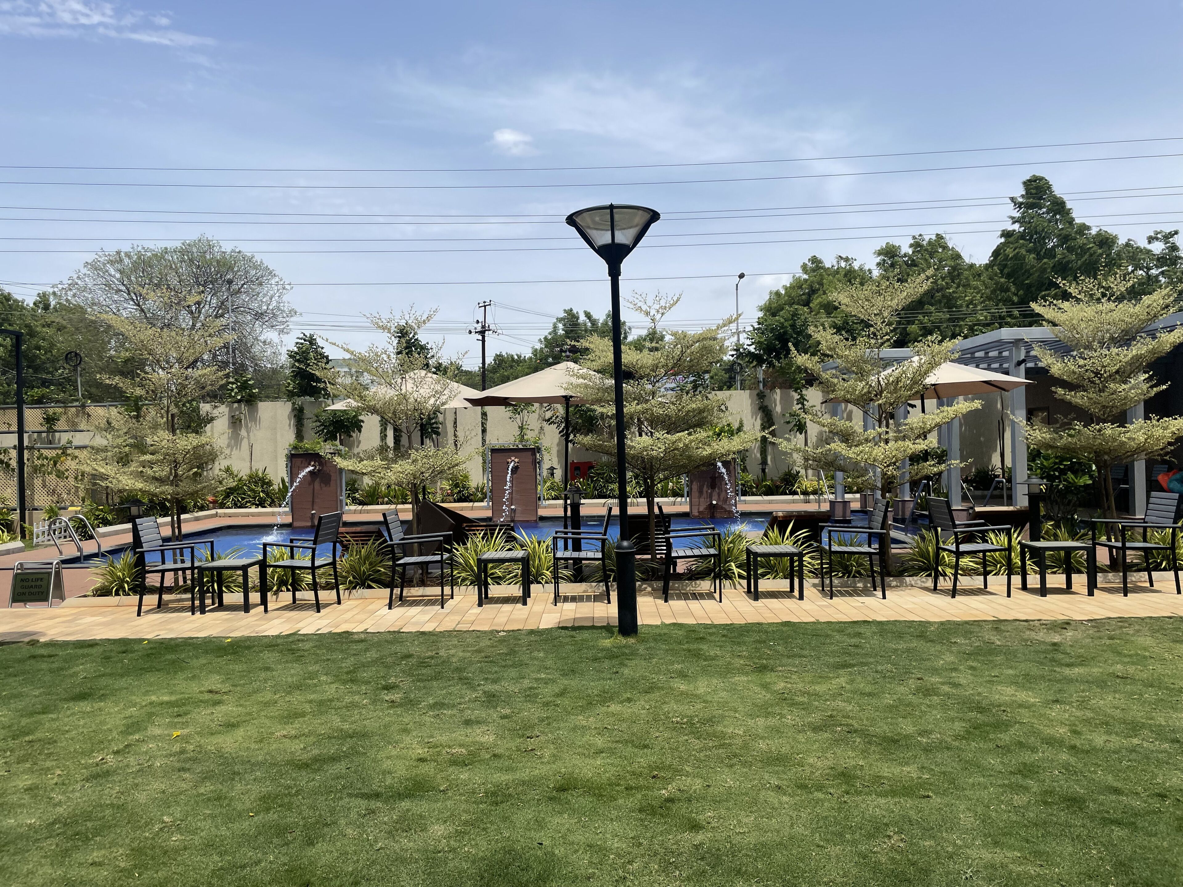 A pool area with lounge chairs and umbrellas, surrounded by greenery and a clear sky.