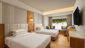 A hotel room with two beds, a desk, a TV, and a window with a view of greenery.