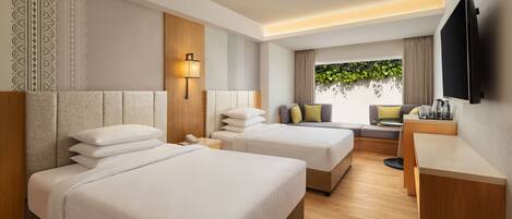A hotel room with two beds, a desk, a TV, and a window with a view of greenery.