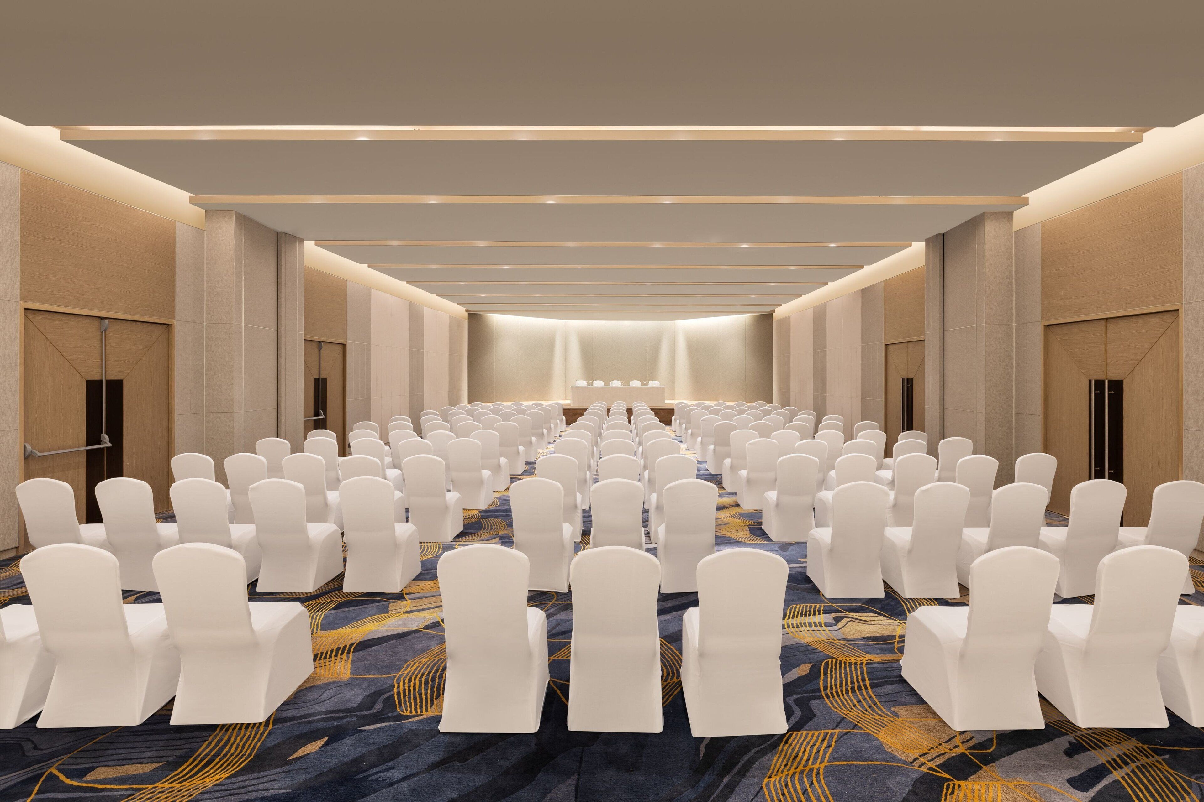 A conference room with rows of white chairs, a patterned carpet, and a stage at the front.