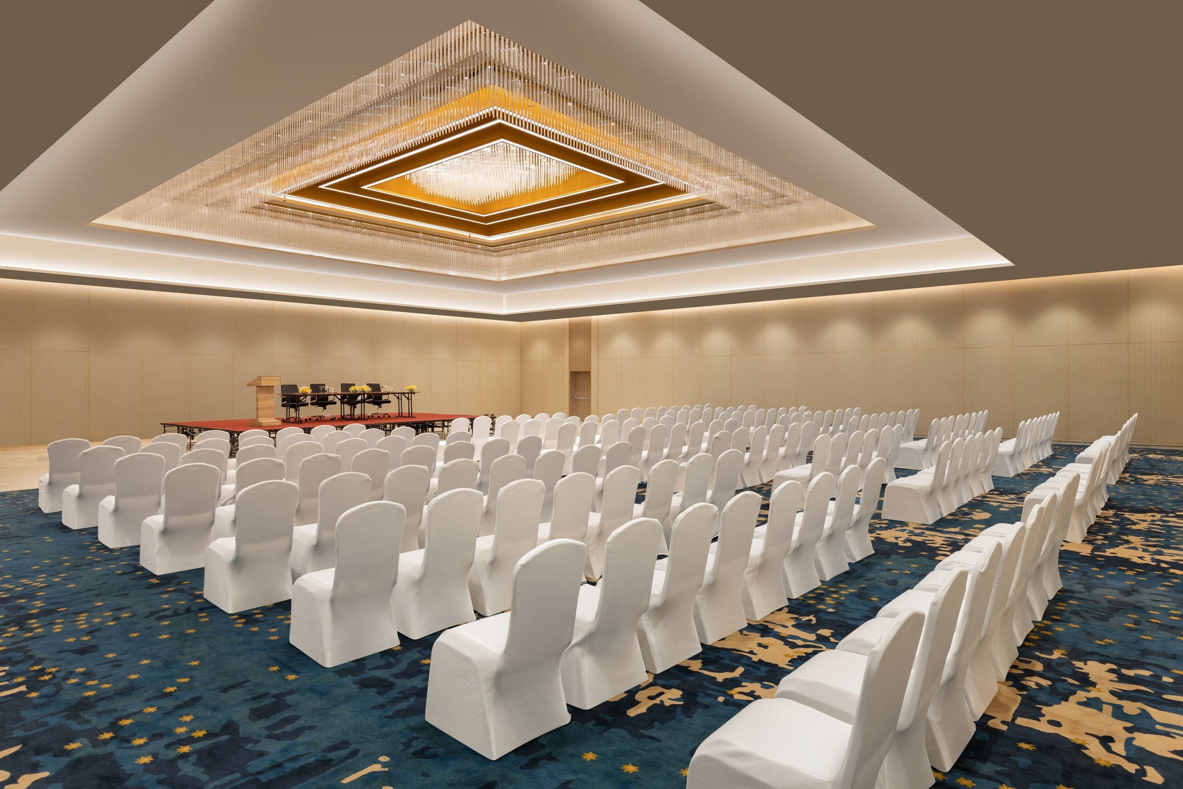 Un salón de conferencias con filas de sillas blancas, una mesa larga y una alfombra estampada.