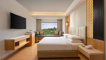 A modern hotel room with a large bed, a flat-screen TV, a desk, and a view of greenery outside.