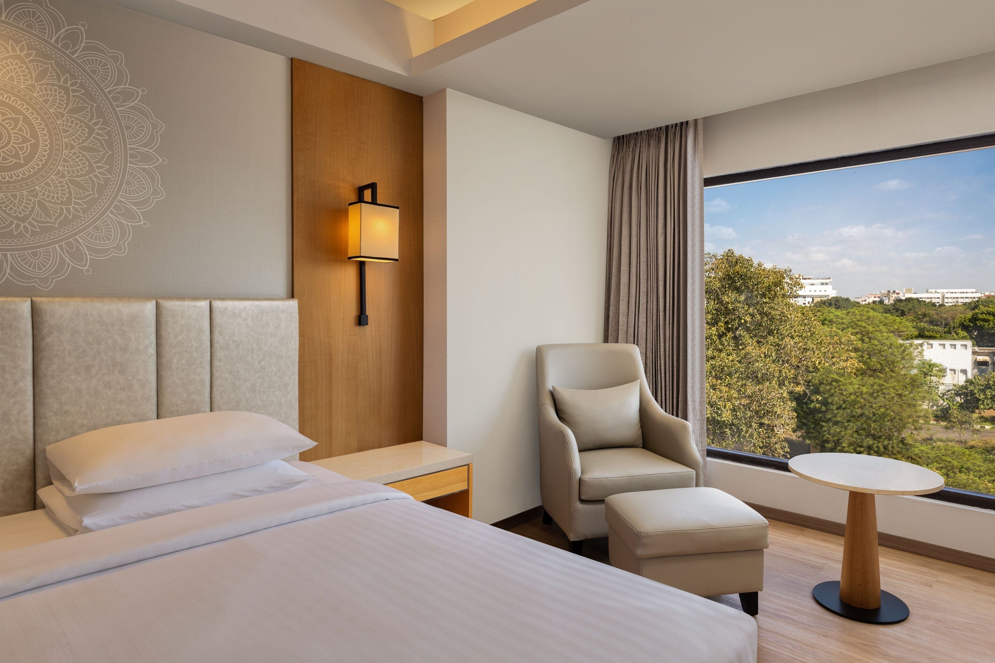 A hotel room with a bed, a chair, a small table, a nightstand, a lamp, and a large window with a view of buildings and trees.