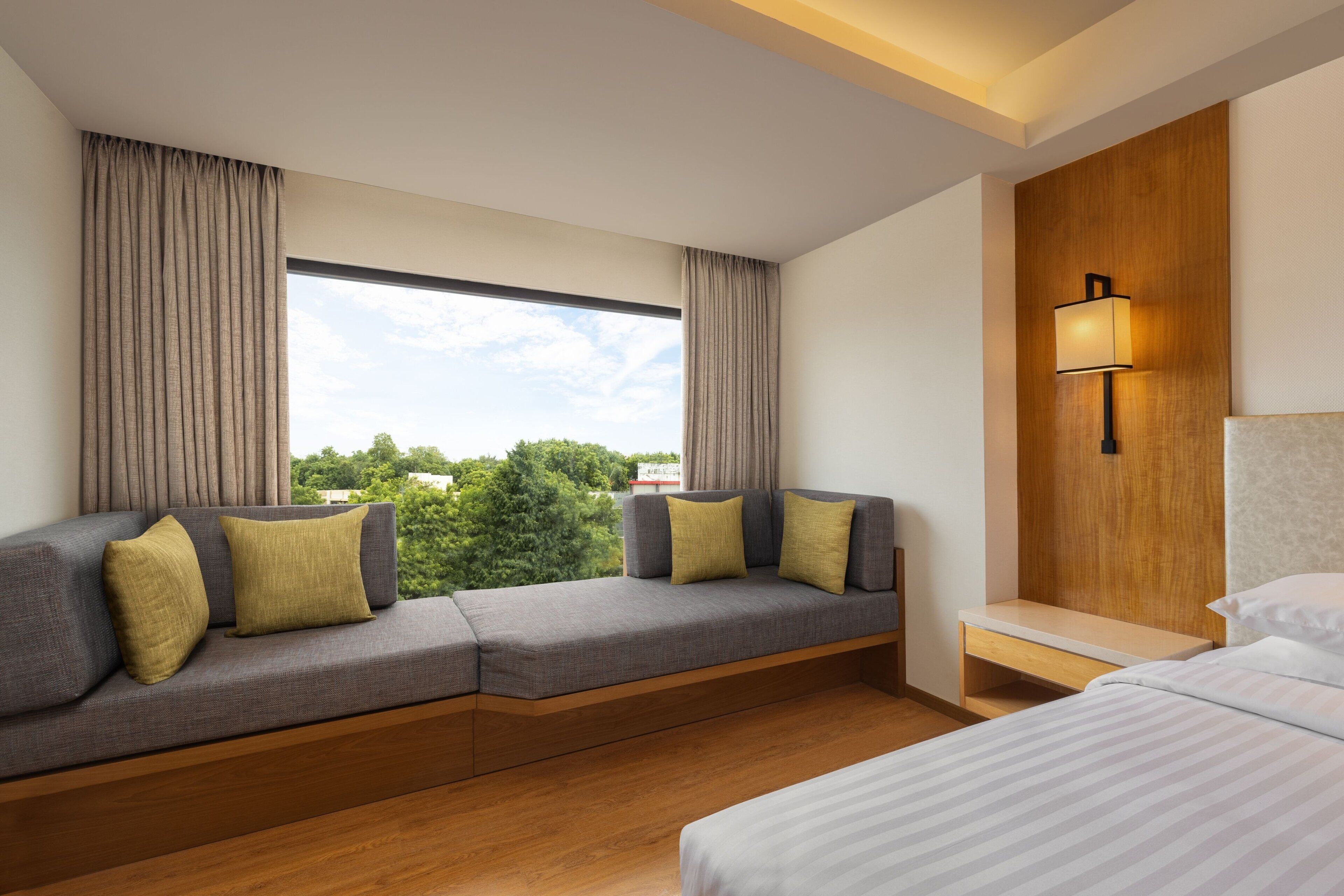 Une chambre d’hôtel moderne avec une grande fenêtre, un canapé avec des coussins, une table de chevet et une lampe.