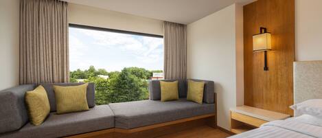 A modern hotel room with a large window, a sofa with cushions, a bedside table, and a lamp.