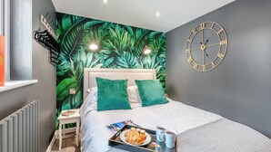 A modern bedroom with a tropical leaf wall mural, a bed with green pillows, a bedside table, and a clock on the wall. - Vitamin Sea Residency (Herne Bay)