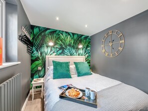 Een moderne slaapkamer met een wanddecoratie van tropische bladeren, een bed met groene kussens, een nachtkastje en een klok aan de muur.