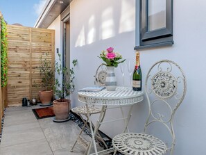 Een klein terras buiten met een ronde tafel, twee stoelen, een bloempot, een vaas met bloemen en een fles champagne.