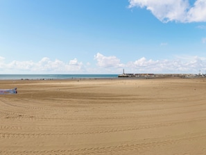 Une vaste plage de sable où l’on peut voir des empreintes, un phare situé au loin et une jetée bordée de bâtiments.