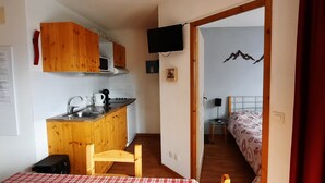 Ett litet kök med träskåp, en mikrovågsugn och ett handfat. I det intilliggande rummet finns ett litet bord med en stol, en röd gardin och en säng med ett mönstrat överkast.
