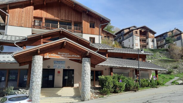 En byggnad i chaletstil med en entré av sten, träbalkonger och en skylt med texten "Residence Le Hamel des Faux d'Orelle".