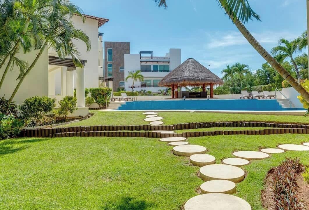 Una piscina con techo de paja, rodeada de césped cuidado y palmeras.