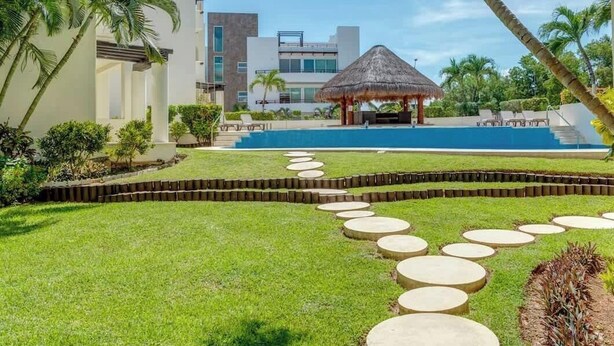 Una piscina con techo de paja, rodeada de césped cuidado y palmeras.