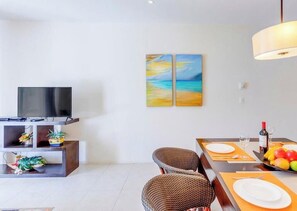 Ein modernes Wohnzimmer mit Flachbildfernseher, zwei Korbstühlen, einem Esstisch mit Obstschale und einem Gemälde mit Strandmotiv.
