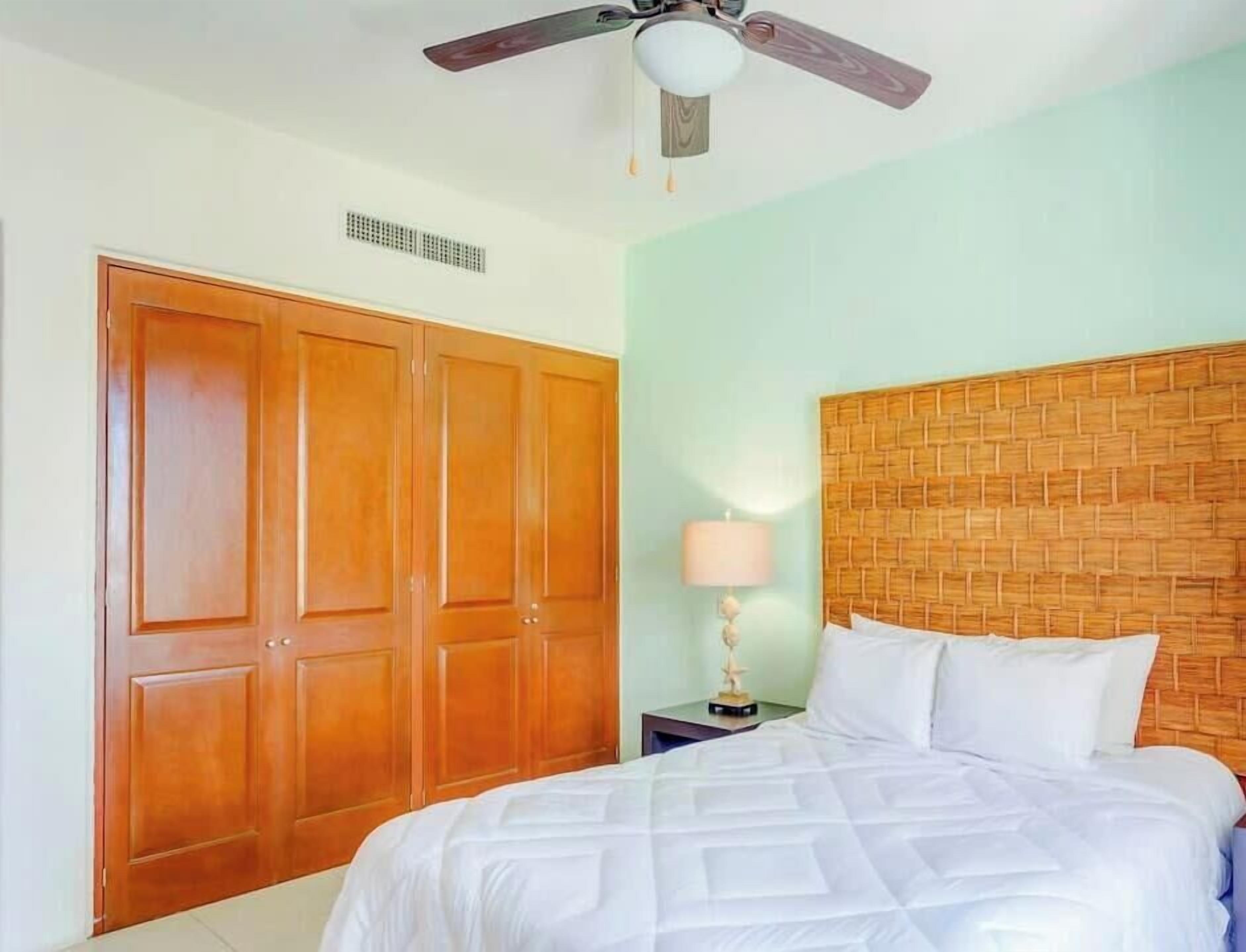 A hotel room with a bed, a wooden wardrobe, a nightstand with a lamp, and a ceiling fan.