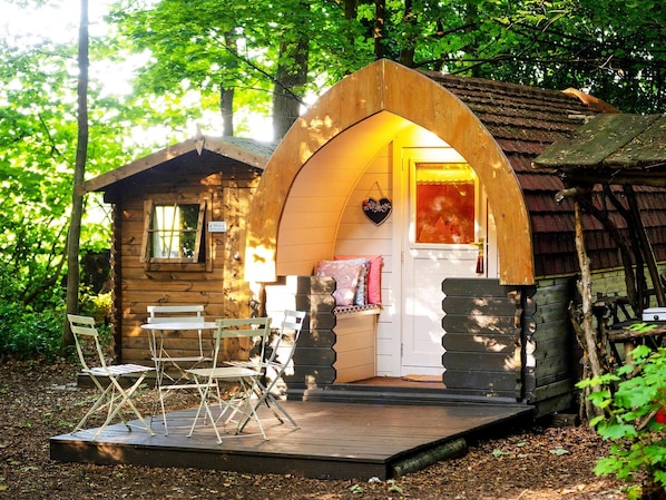 A rustic wooden cabin with an arched entrance, a small porch with folding chairs, and a heart-shaped decoration on the wall.