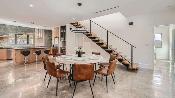 Una cocina moderna con una isla central, un comedor con mesa y sillas redondas, y una escalera con barandilla de vidrio.