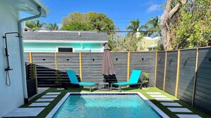 Una piscina rectangular con dos sillas reclinables azules y una pequeña mesa, rodeada por una valla de madera y un césped.