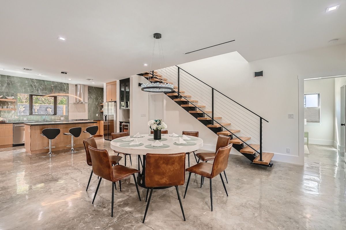 Eine moderne Küche mit Marmorfliesenspiegel, ein Essbereich mit rundem Tisch und Stühlen sowie eine Treppe mit Glastrennwand.