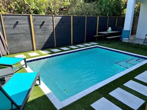 Una piscina rectangular con bordeado de azulejos, rodeada por un césped con piedras para pisar, y dos sillas reclinables azules.