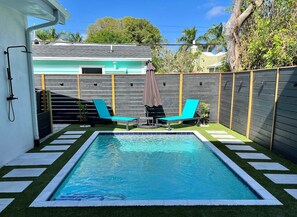 A rectangular swimming pool with two lounge chairs and a small table, surrounded by a wooden fence and a grassy area.