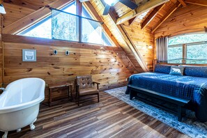 A wooden cabin bedroom with a sloped ceiling, a large window, a bathtub, a bed, and a chair. - 3BR Cabin: Hot Tub, Game Room, Near Strip, Porches (Pigeon Forge)