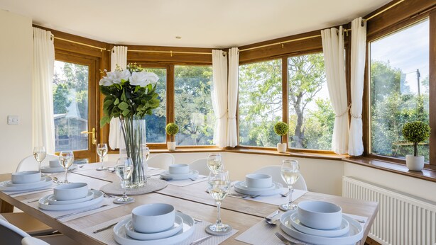 A dining room with a long table set for a meal, large windows with curtains, and a view of greenery outside. - Stunning 5-Bedroom Home in Truro (Truro)