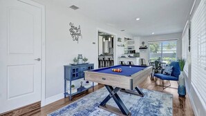 Un salon moderne avec un billard, un canapé bleu et un coin cuisine visible à travers une porte ouverte.
