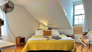 A bedroom with a sloped ceiling, a bed with a yellow bedspread, a bedside table, a nightstand, a fan, and a window. - Townhouse Apt 2 w Parking in Downtown Portsmouth (Portsmouth)